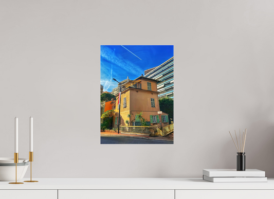 Square framed photo print under acrylic glass showing a yellow building, green shutters, and bright blue sky.