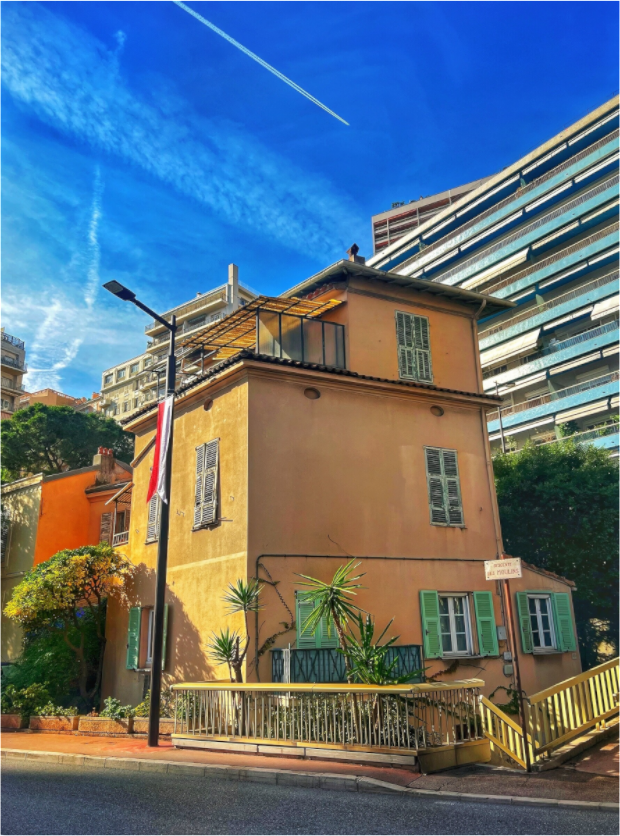 Square framed photo print under acrylic glass showing a yellow building, green shutters, blue sky.