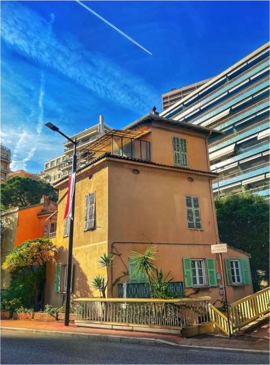 Square framed photo print under acrylic glass showing a yellow building, green shutters, blue sky.