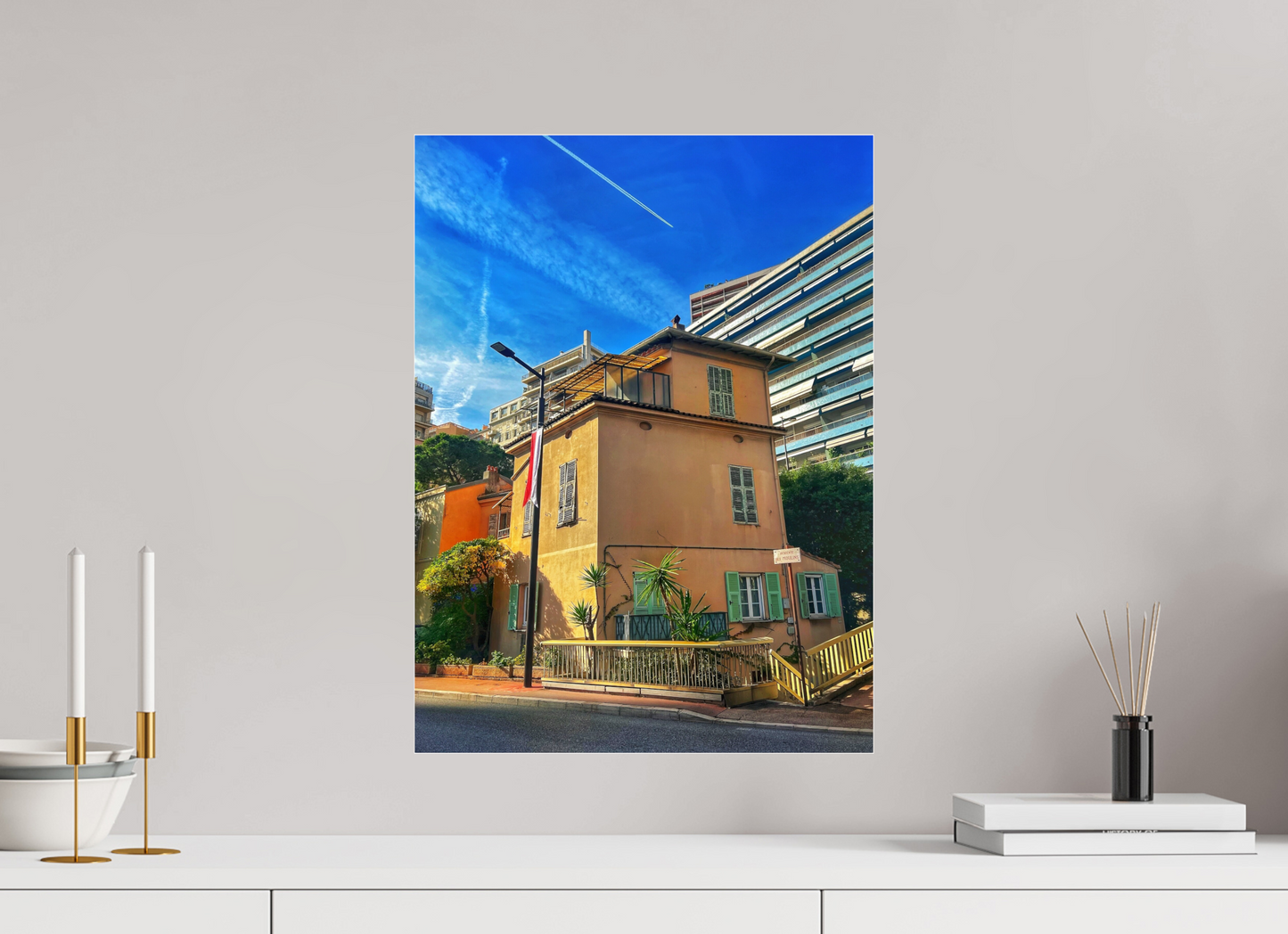 Square framed photo print under acrylic glass featuring a yellow house, plants along the walkway, blue sky.