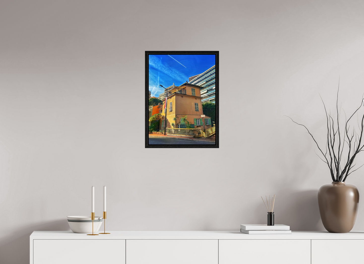 Framed street scene photo print in black floater frame, showing a yellow house facade and greenery.